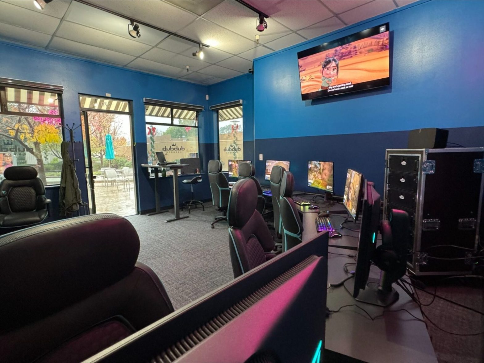 The Greenbrae Esports Gaming Center features a dynamic gaming room with multiple computers and comfy chairs facing sleek monitors. A TV on the blue wall immerses players in video game action, while large windows reveal a scenic patio and lush trees outside.