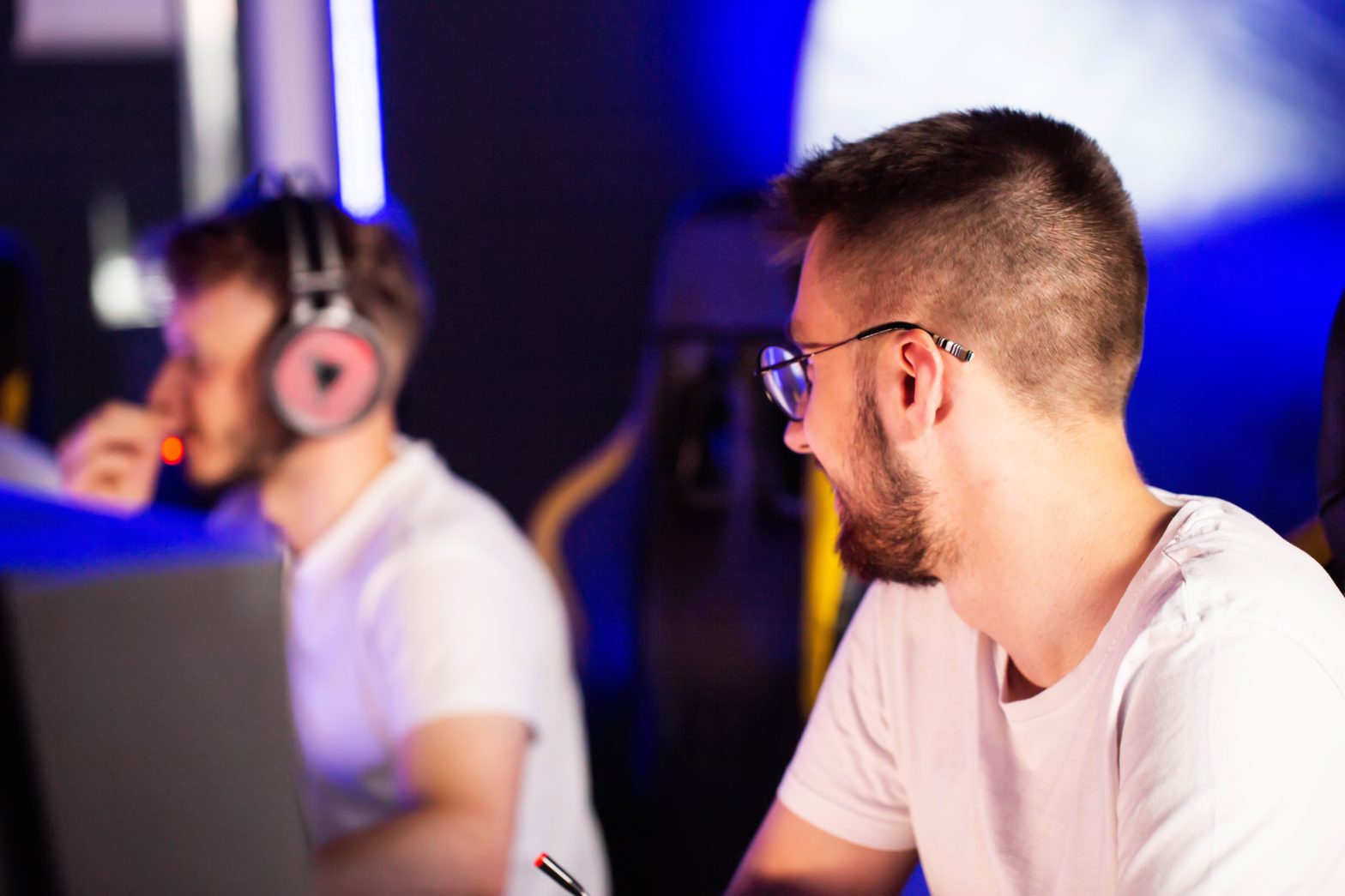 Two men in white shirts sit at computers; one wears headphones and faces a screen, while the other, in glasses, looks to the side.