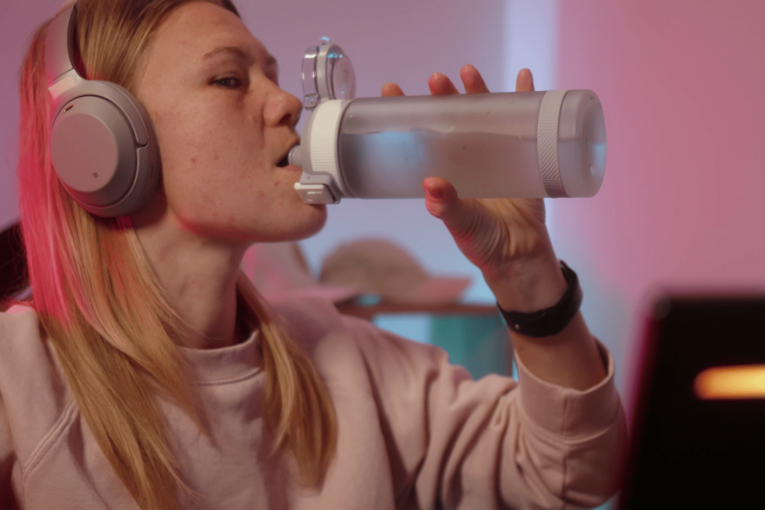 A person wearing headphones and a sweatshirt drinks from a water bottle while sitting indoors.