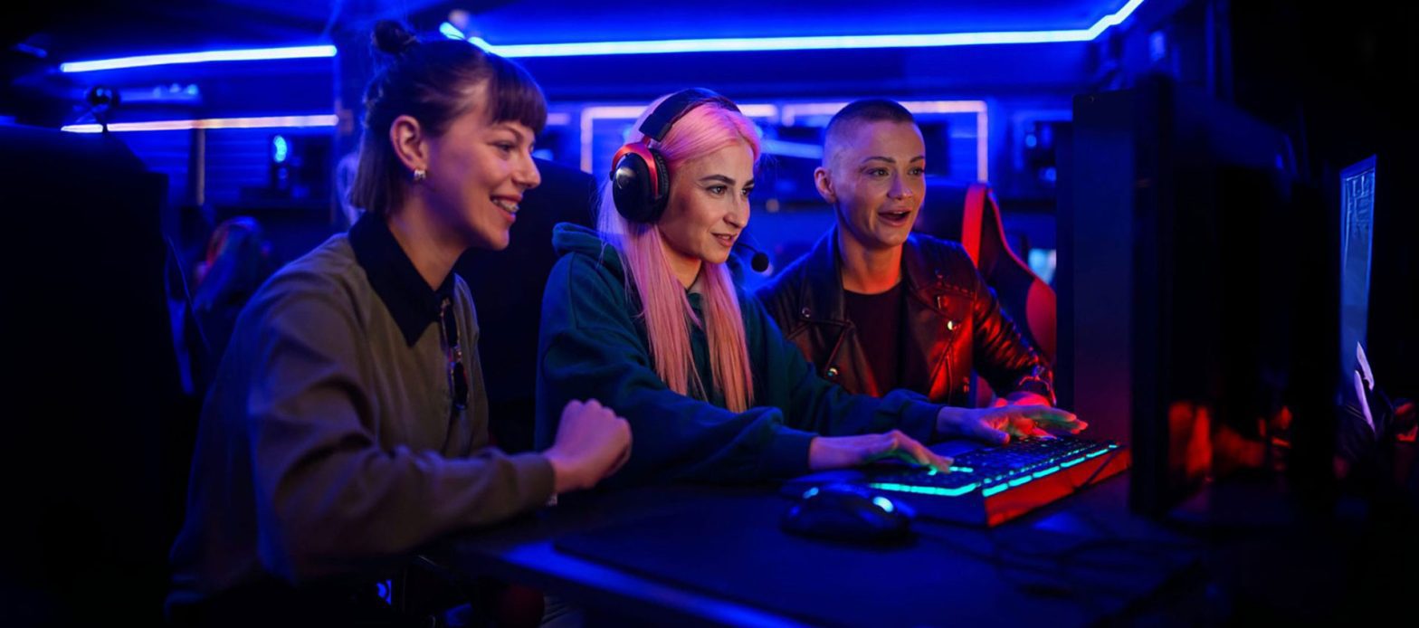 Three people sit side by side in a neon-lit room, focused on a computer screen while playing a game together.
