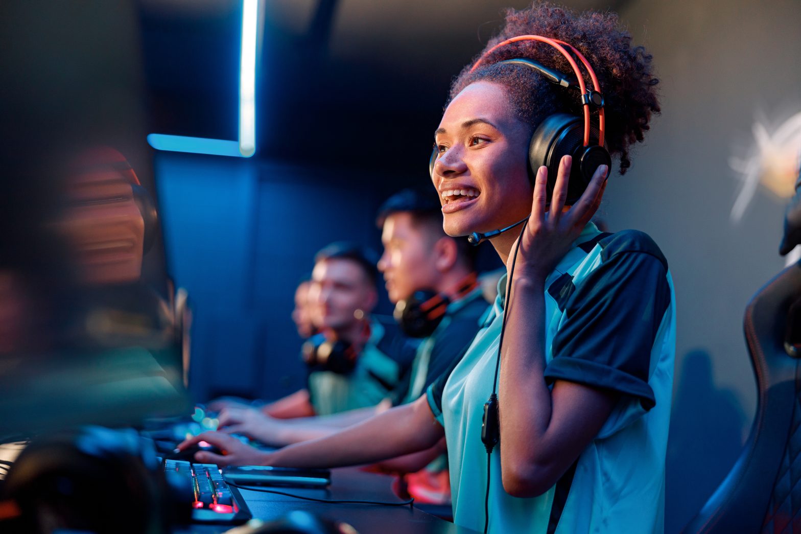 A young woman wearing headphones and a gaming jersey smiles while playing at a computer in a row with other gamers.